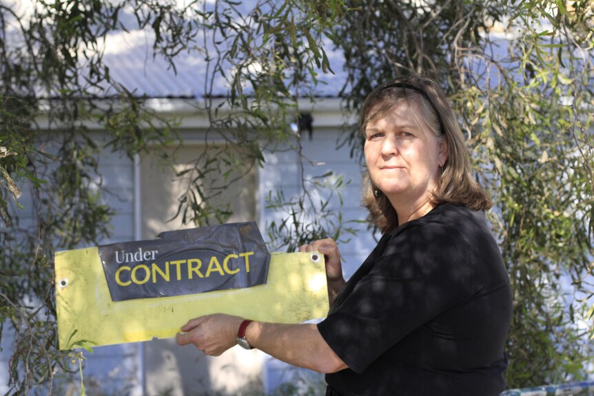 A lady holds a destroyed real estate sign that says 'Under Contract' in front of an old blue house