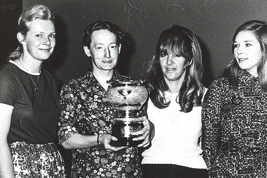 Black-and-white photograph of four smiling women, with one holding a trophy.