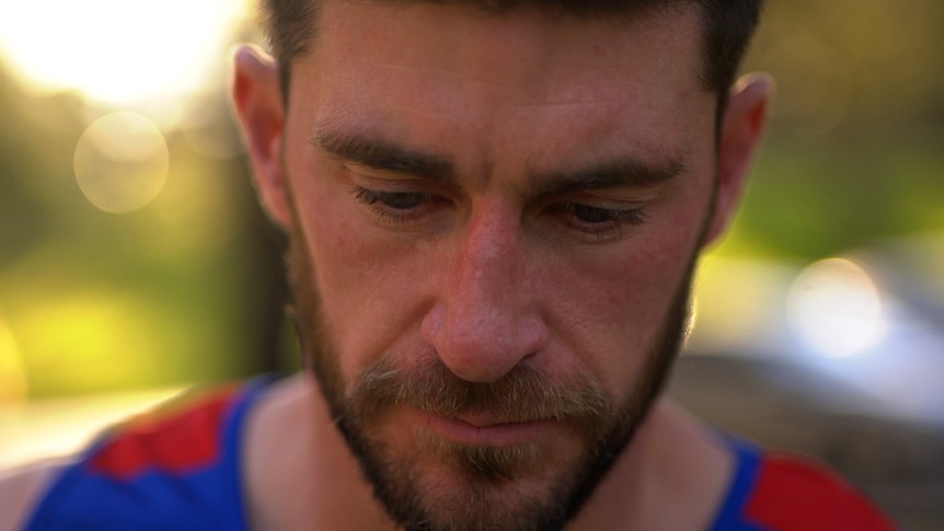 A close up of a man's face, his eyes look downwards. He is wearing a blue and red sports singlet.