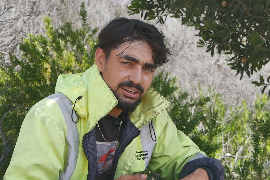 He sits under a tree on the island, in a high-vis jacket, looking at the camera, fairly close up