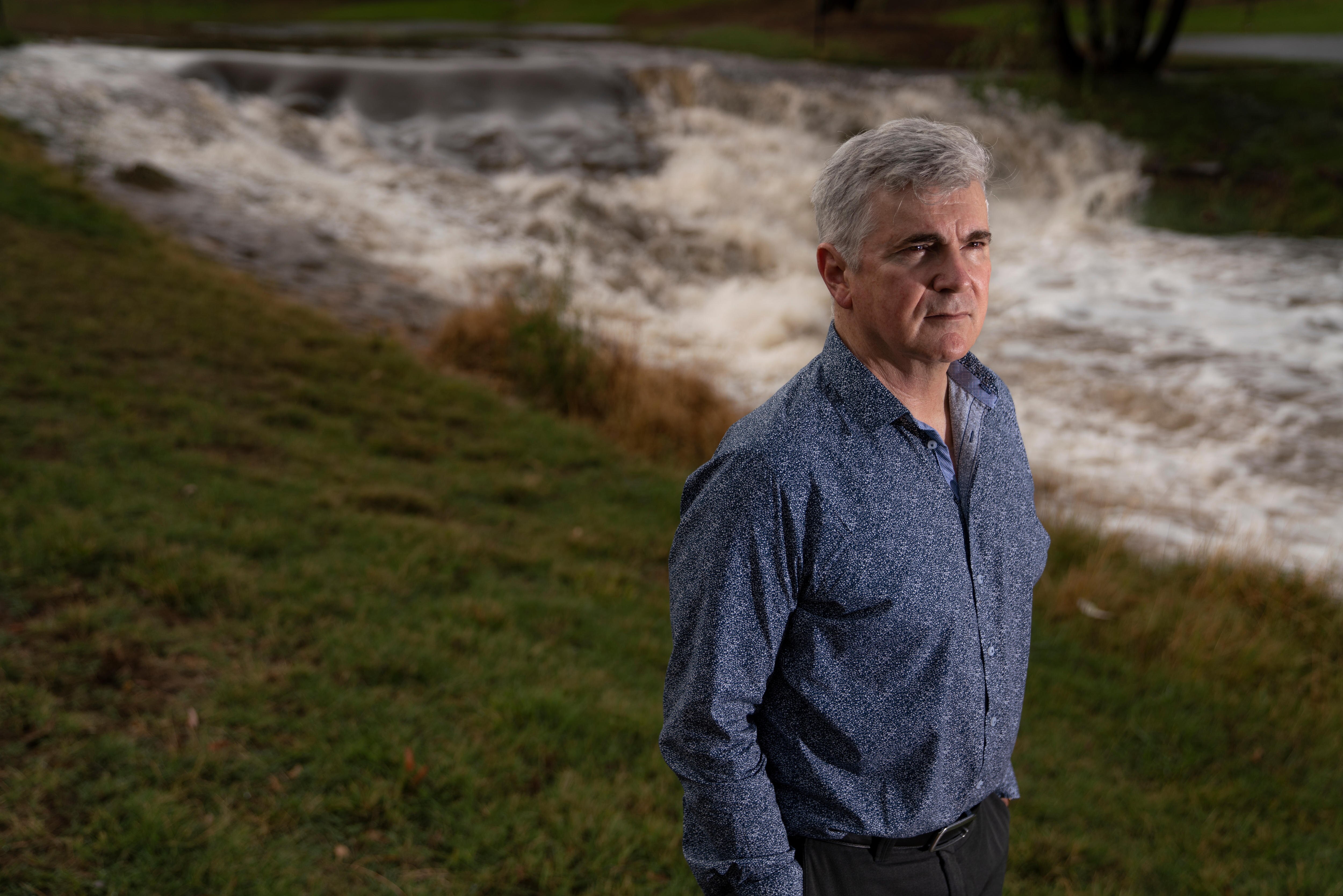 Professor Mark Howden stands almost side-on in front of a rushing river