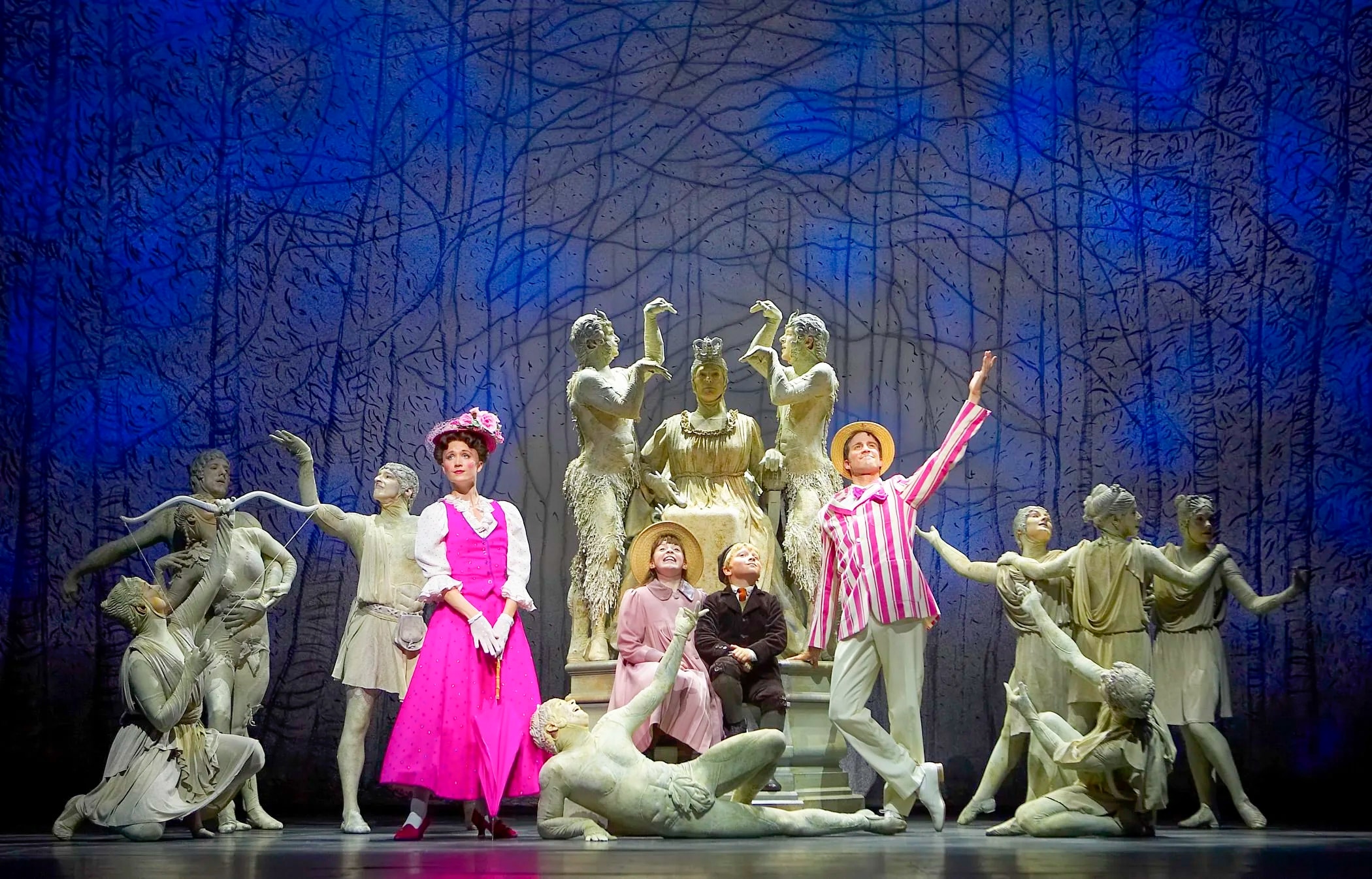 An image of a stage performance of Mary Poppins in the Park, with Mary, Burt, Jane and Michael, and several grey statues.