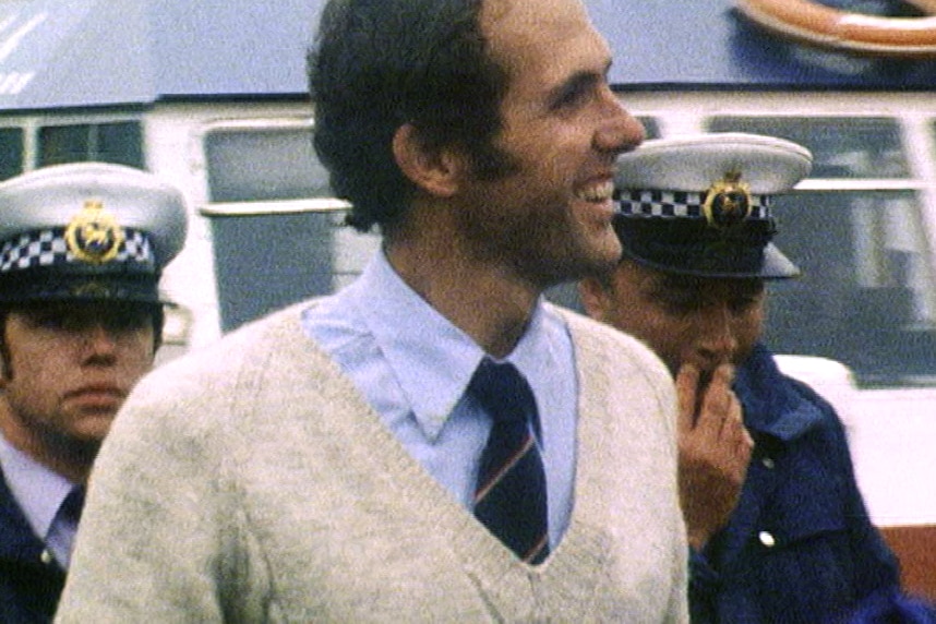 A man in a sweater smiling as he is lead away by two police officers.