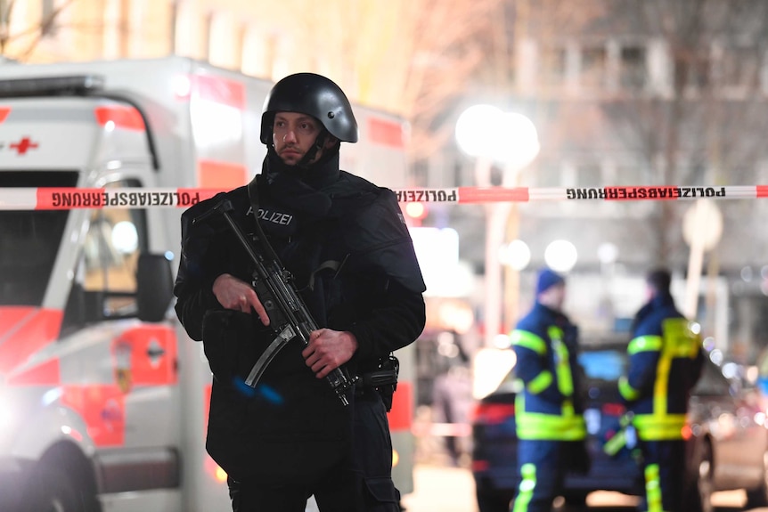 A police officer dressed entirely in black holds a large automatic weapon. An ambulance is behind him.