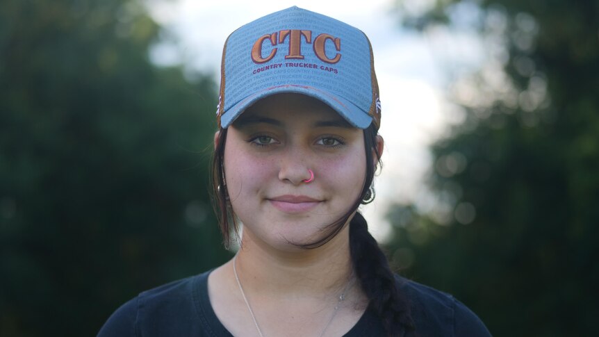 A young girl wearing a blue trucker cap with the initials C T C smiles slightly at the camera.