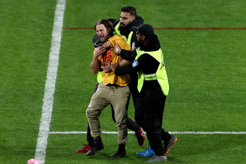 Two security guards escort a man off a football pitch. One has his arm around the man's neck