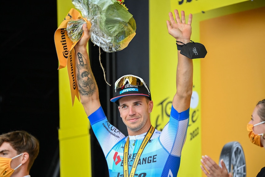 Dylan Groenewegen stands on the podium with hands up, wearing a medal and holding flowers