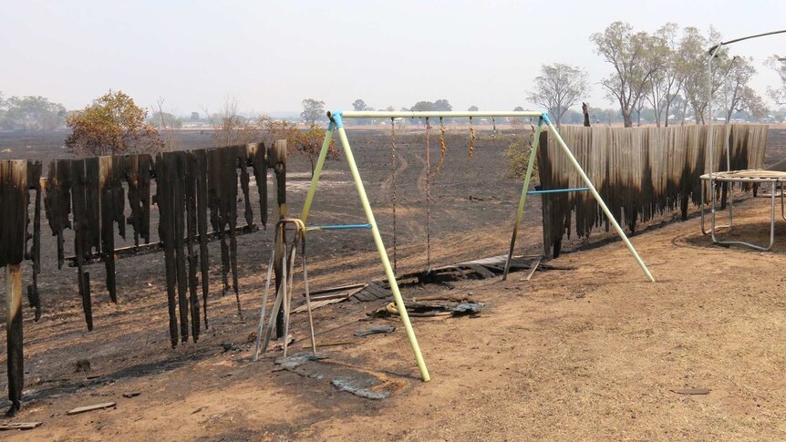 A wooden fence and children's swing set damaged by fire, beyond it is scorched black ground.
