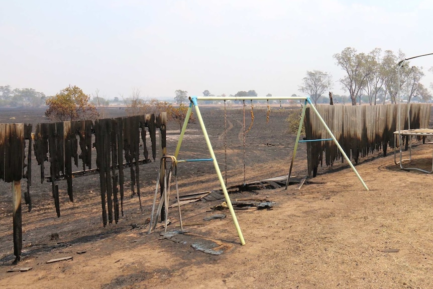 A wooden fence and children's swing set damaged by fire, beyond it is scorched black ground.