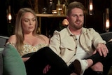 A man and a woman sit next to each other on a confessional couch on a reality show. The woman looks unhappy as the man talks.