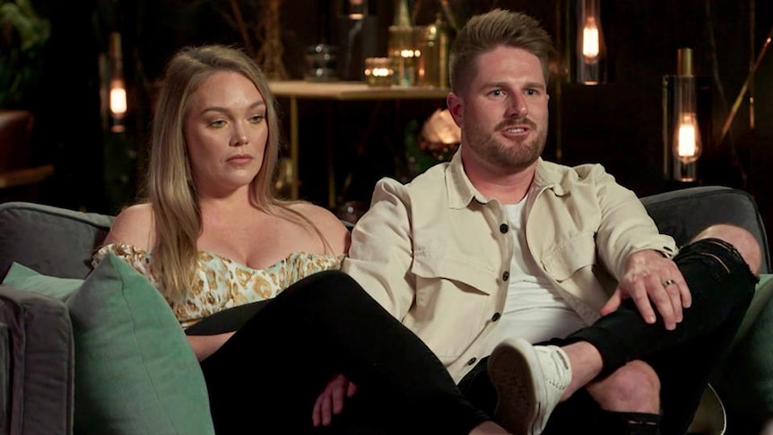 A man and a woman sit next to each other on a confessional couch on a reality show. The woman looks unhappy as the man talks.