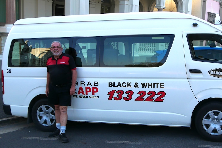 A taxi driver wearing a black and red shirt and shorts standing outside a taxi van.