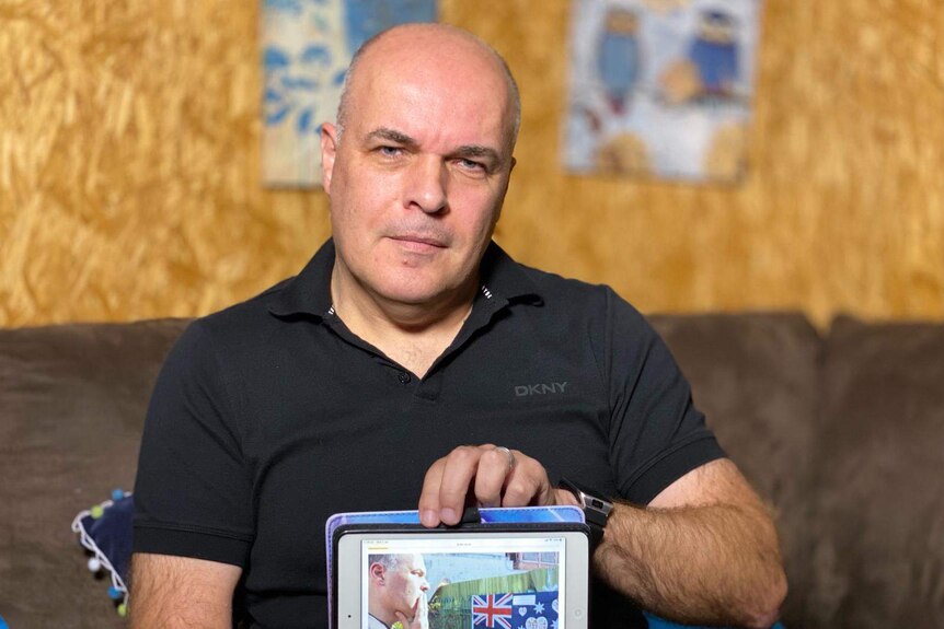 A bald man wearing a black shirt holds a photo of himself at a memorial as he sits on a brown couch.