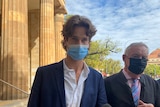 A young man wearing a face mask stands in front of a court building, his lawyer beside him