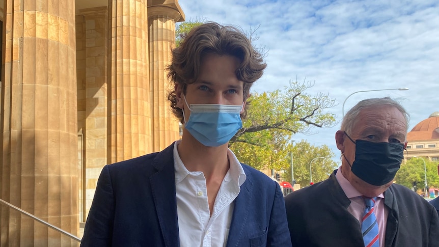 A young man wearing a face mask stands in front of a court building, his lawyer beside him