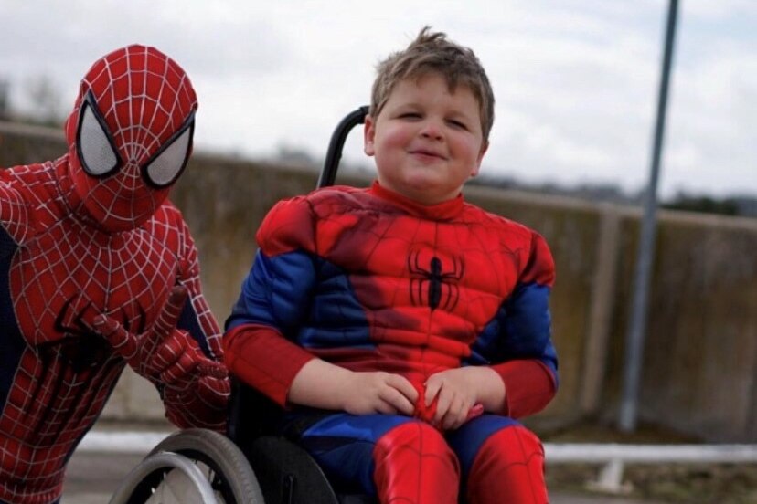 A young boy in a superhero suit, sitting in a wheelchair next to Spiderman.
