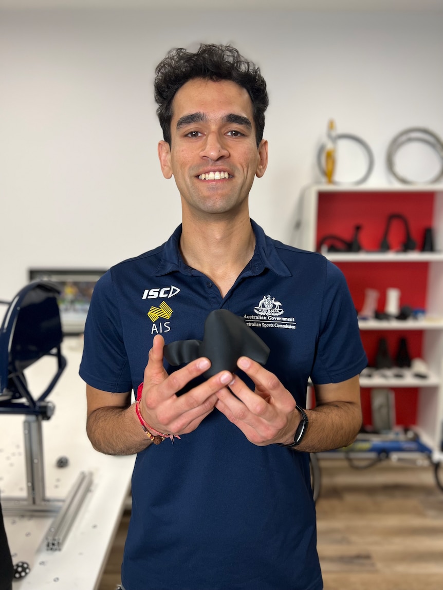 A man wearing an AIS shirt with an 'Australian Government Australian Sports Commission" logo stands holding a black glove.