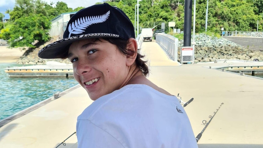 A young teenage boy sits on a pier with fishing rods around him, smiling.