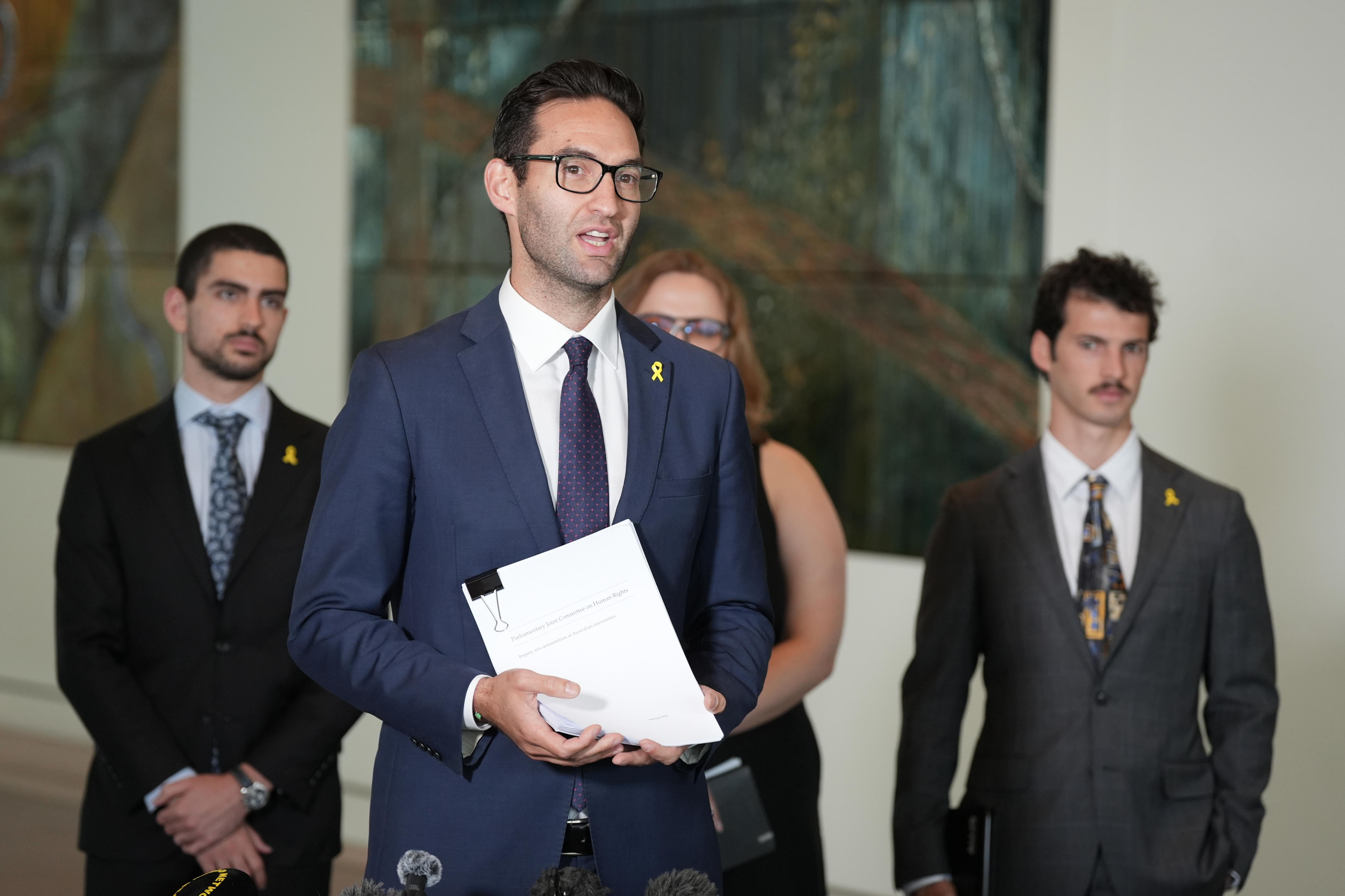 Josh Burns sostiene el Informe Parlamentario en el Parlamento. Hay tres estudiantes detrás de él.