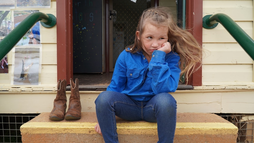 A little girl sits on a step next to a pair of work boots, cupping her chin in her hand and looking pensive.