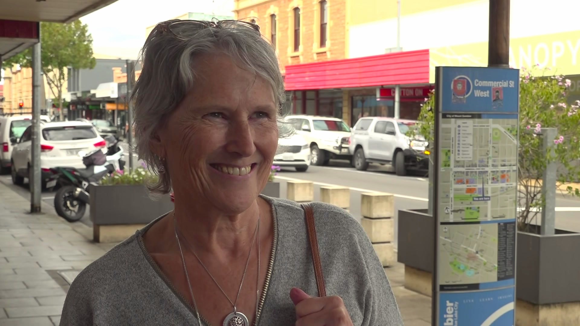 Μια ηλικιωμένη γυναίκα με κοντά μαλλιά που φορά ένα γκρι πουκάμισο και κρατά το καφέ λουράκι μιας τσάντας στέκεται σε ένα πολυσύχναστο μονοπάτι.