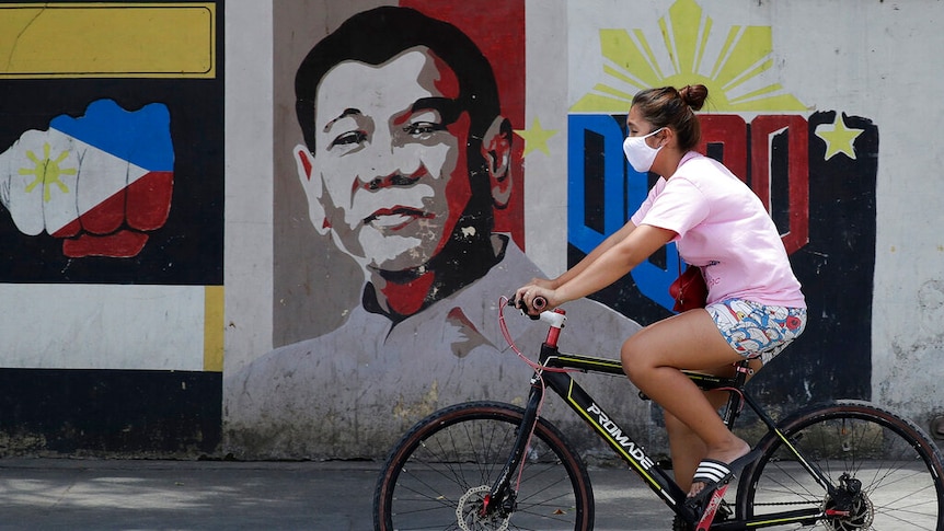 A woman wearing a protective mask rides her bicycle past an image of Philippine President Rodrigo Duterte.