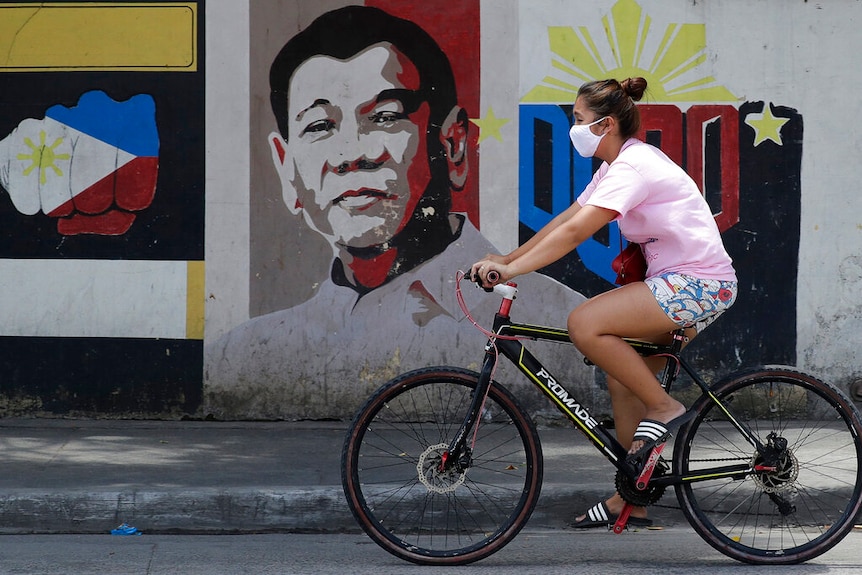 A woman wearing a protective mask rides her bicycle past an image of Philippine President Rodrigo Duterte.