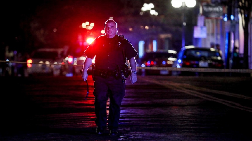 A police officer is seen walking with red lights behind him. He is walking towards the camera with a neutral expression.
