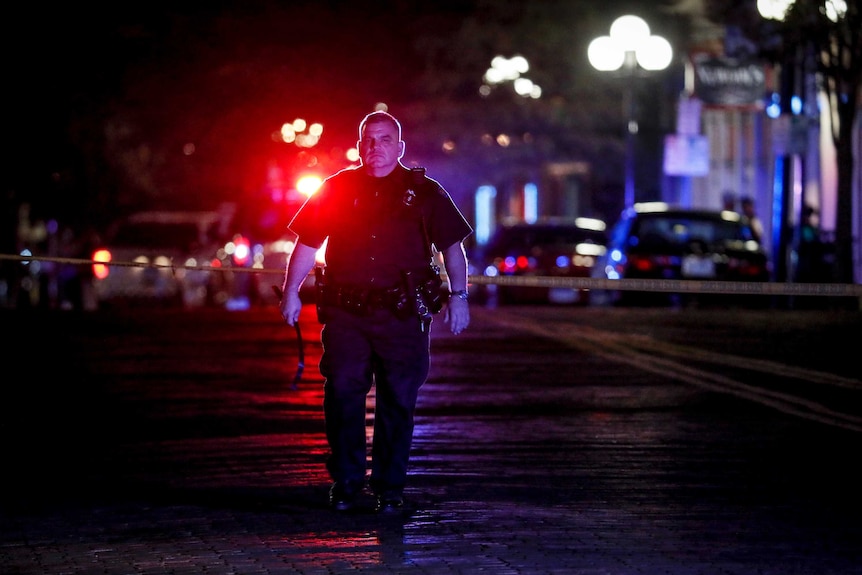 A police officer is seen walking with red lights behind him. He is walking towards the camera with a neutral expression.