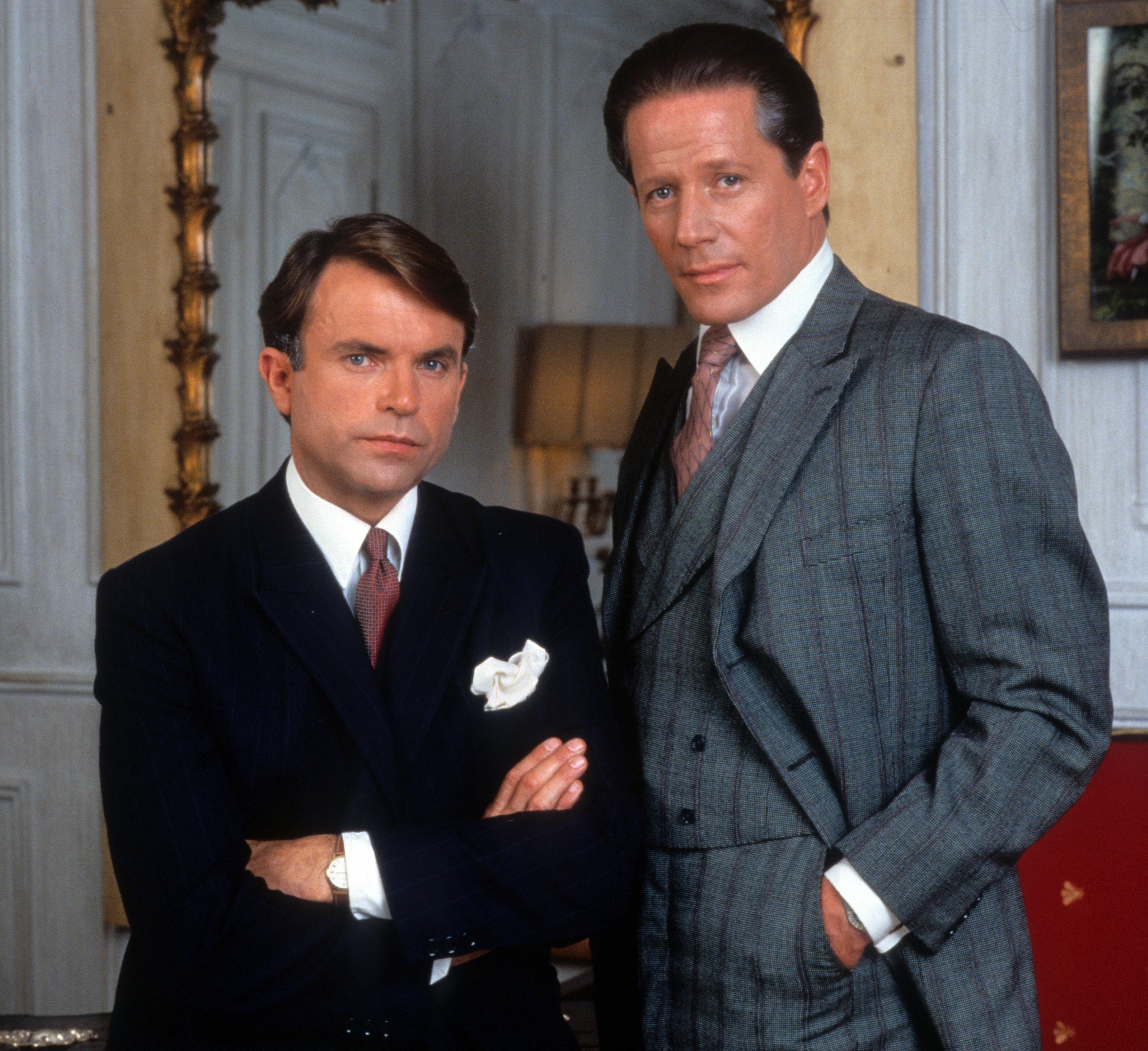 Two men in sharp suits and ties look serious. One man seated has his arms crossed while the other stands with hands in pockets 