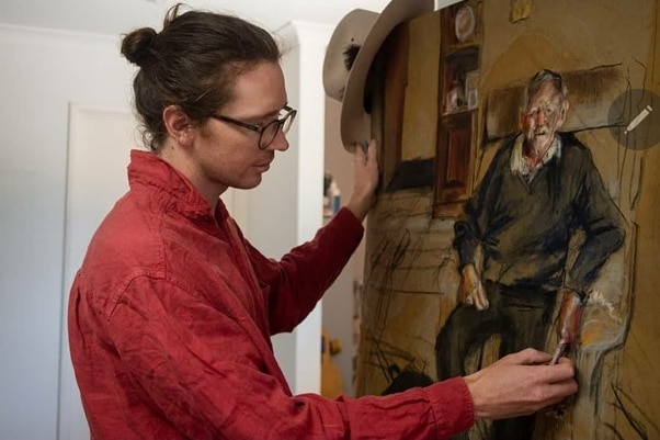 Man with red shirt and glasses uses chalk to draw a picture of an older man in a chair. 