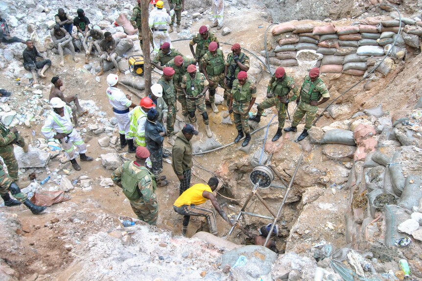 Rescuers in army uniform and red helmets stand around a rocky area. 