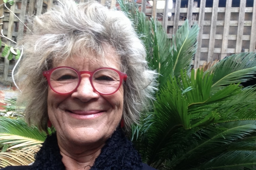 Dr Green is smiling. She has grey hair and red-framed glasses and there is a big green plant behind her.