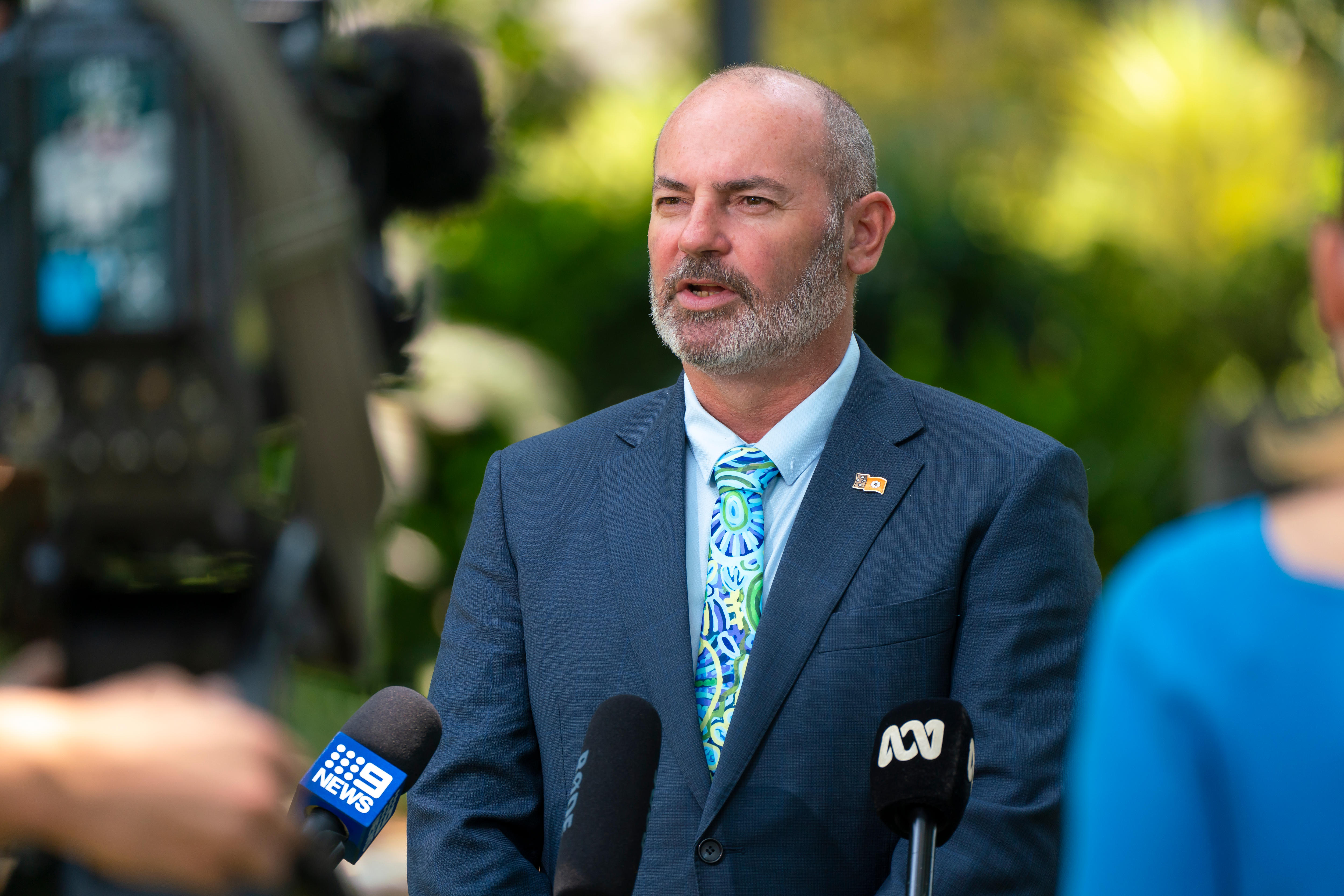 A balding man in a jacket and tie stands at a media microphone