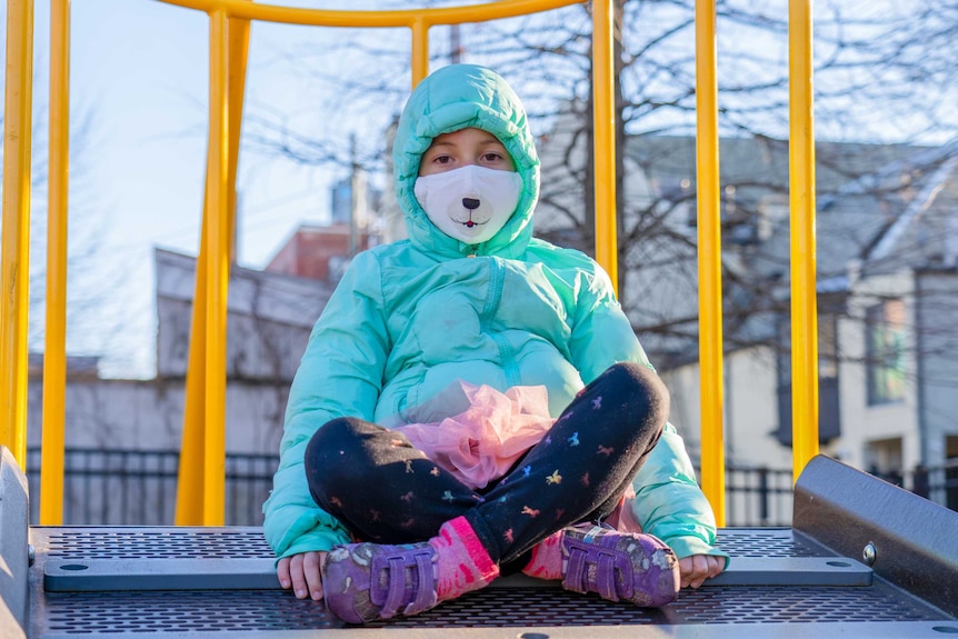 A little girl sits cross legged on a jungle gym while wearing a green parker and face mask