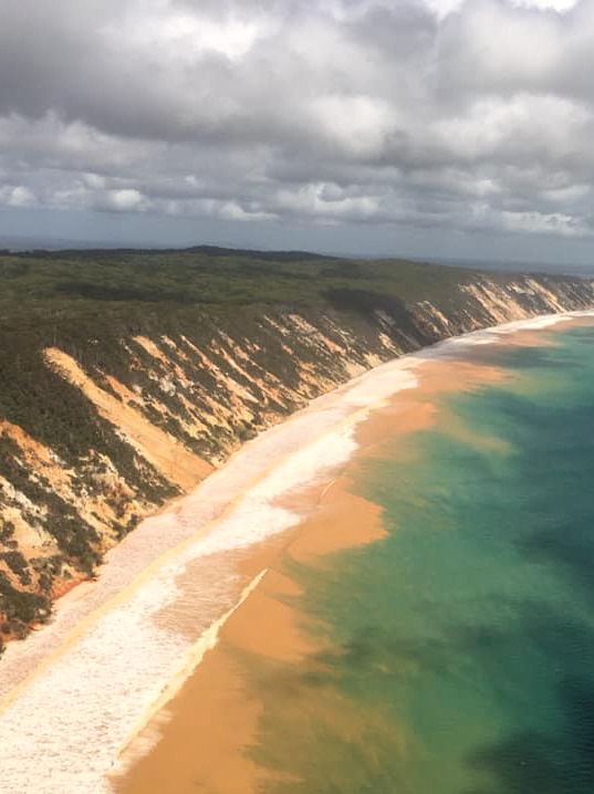 An aerial photo shows waves washing sand from Rainbow Beach into the ocean.