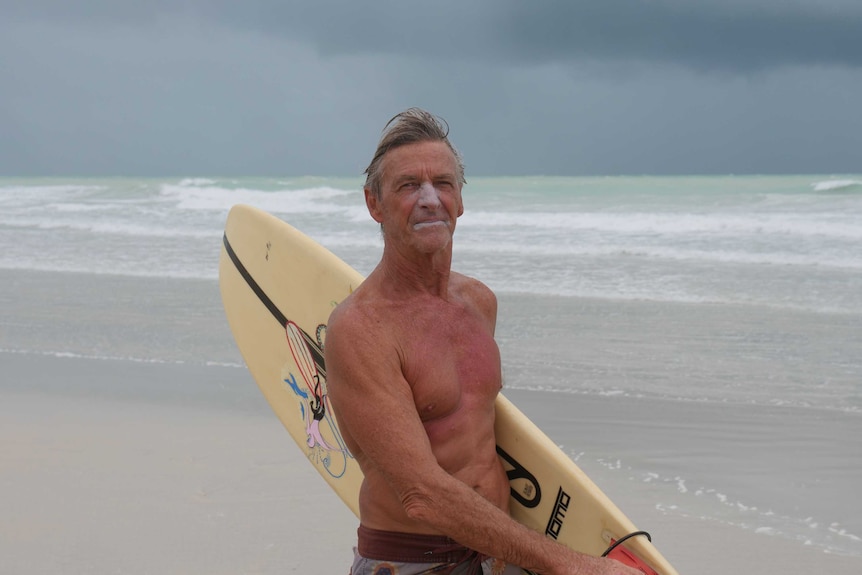 A man with a surfboard walks along a beach with dark clouds in the background.