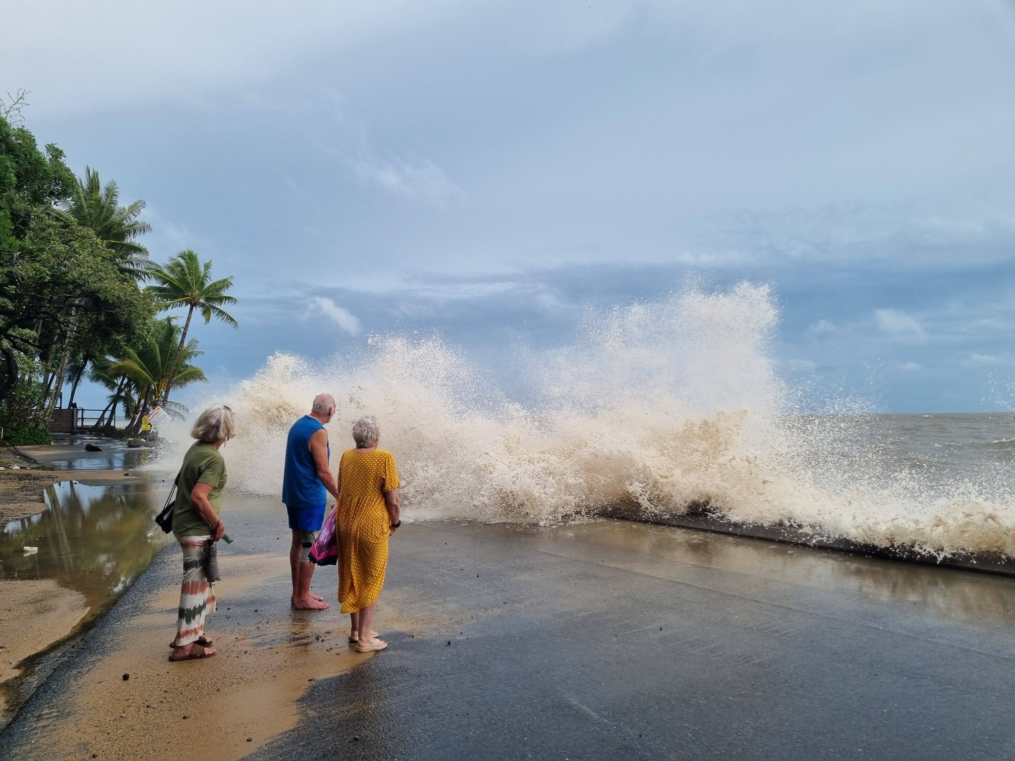 People stand on a beachside path as a wave splashes in front of them.