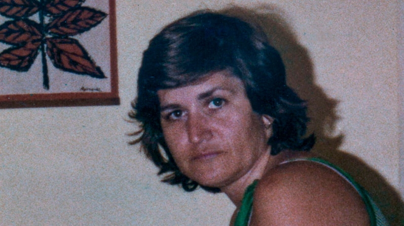 Middle-aged woman with brown hair stares into camera, standing in front of wall.