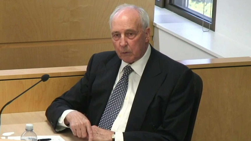 A man wearing a suit speaking at a desk with a microphone