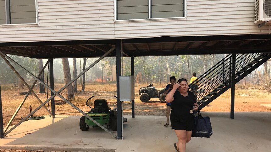 Two people walk from their house, one holding a bag. A bushfire can be seen behind the house.