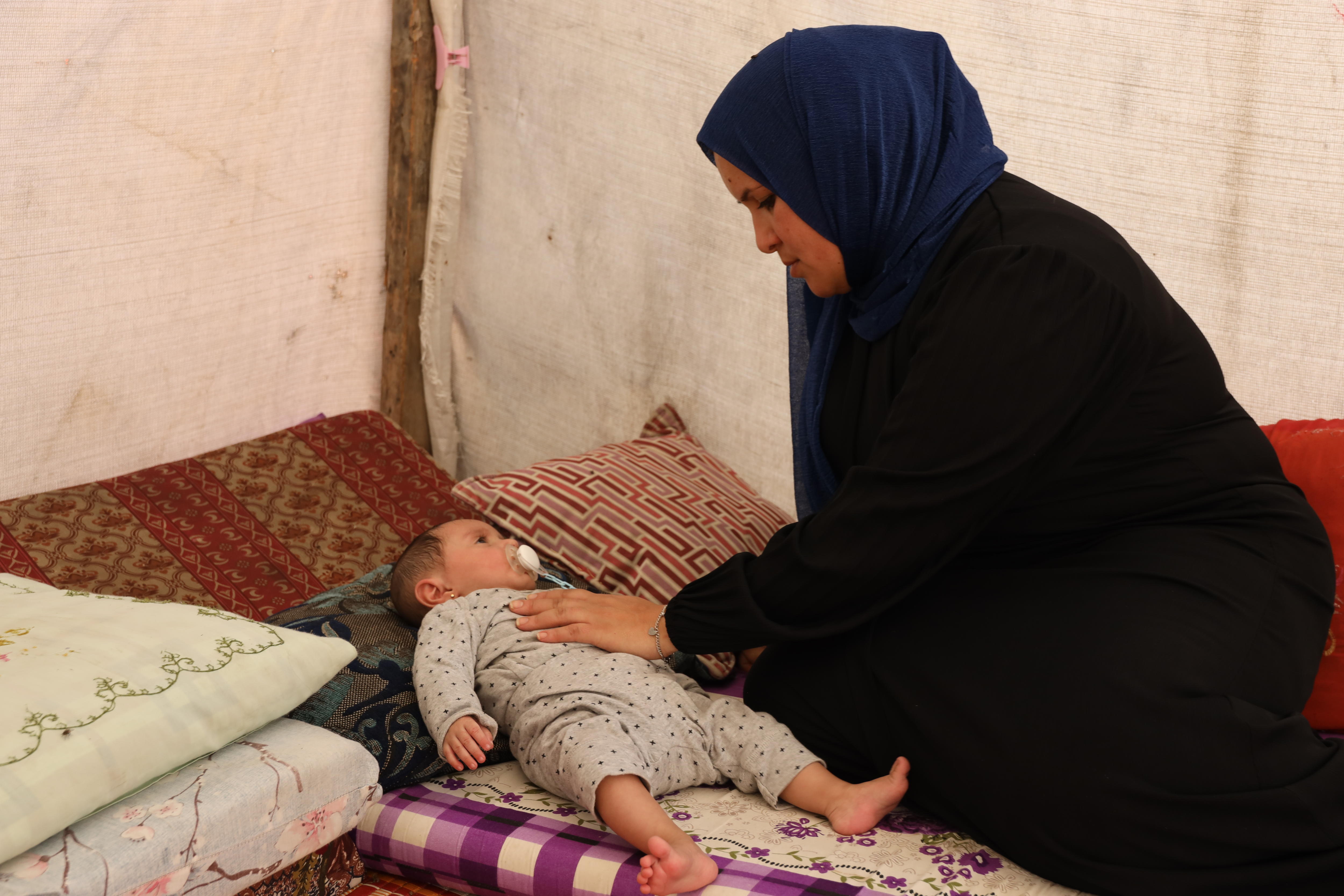 A woman in black clothing and navy hijab looks down at her baby daughter and places a soothing hand on her chest.