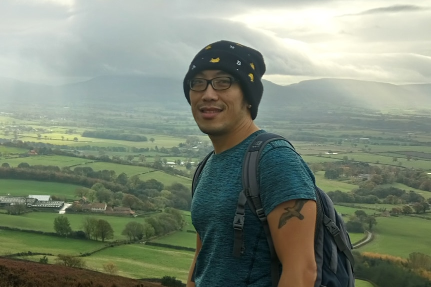 A man stands wearing a backpack overlooking green fields in a rural area