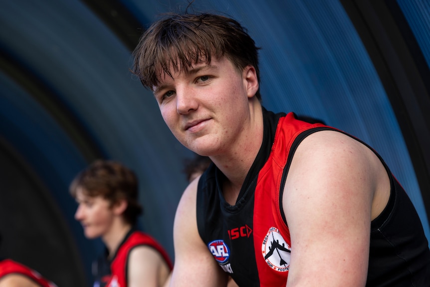 A young man in red and black footy singlet grins at the camera
