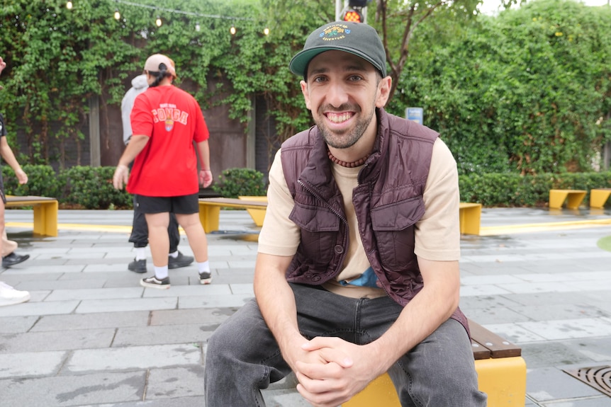 A man in a hat and brown vest smiles while seated in a public square
