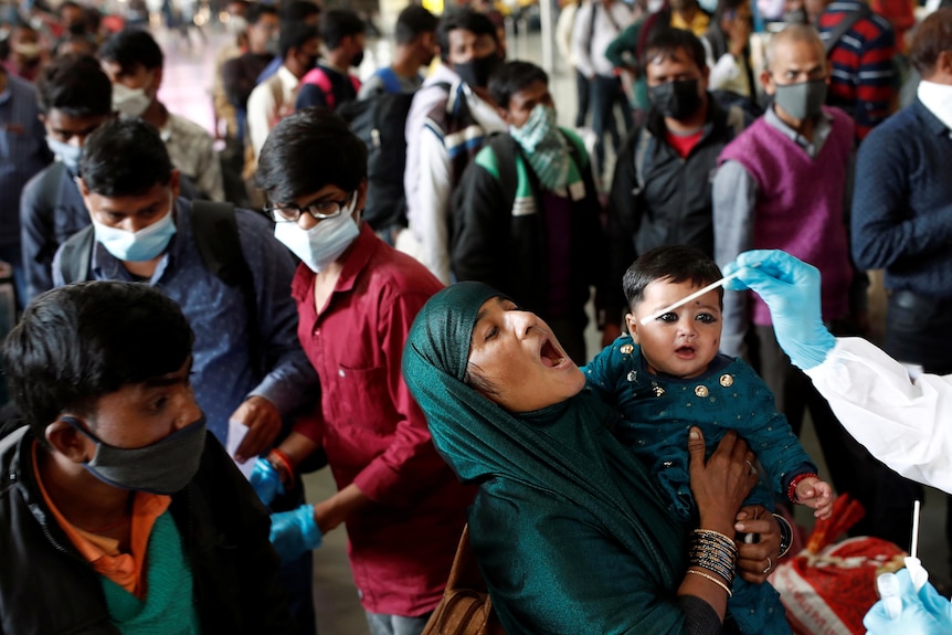 A woman stands in a crowd with her baby in her arms, her mouth open ready to be swabbed by healthcare worker