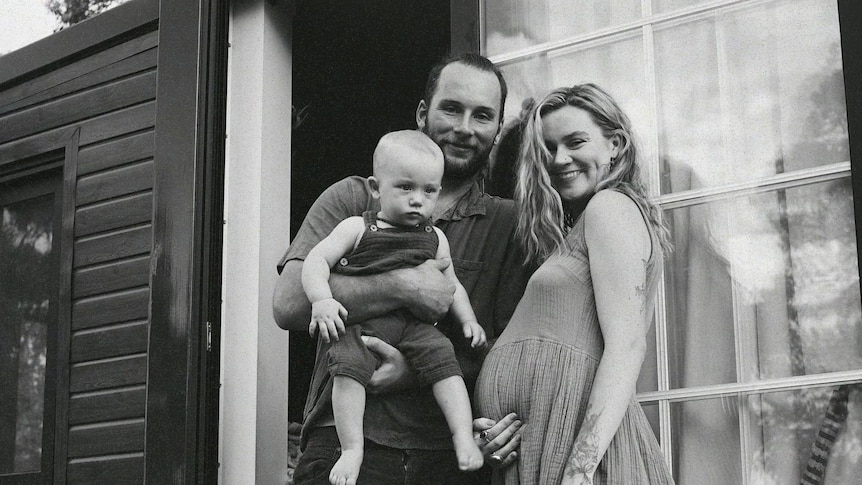 A smiling pregnant woman and her partner, who holds a baby, stand in front of a tiny home.