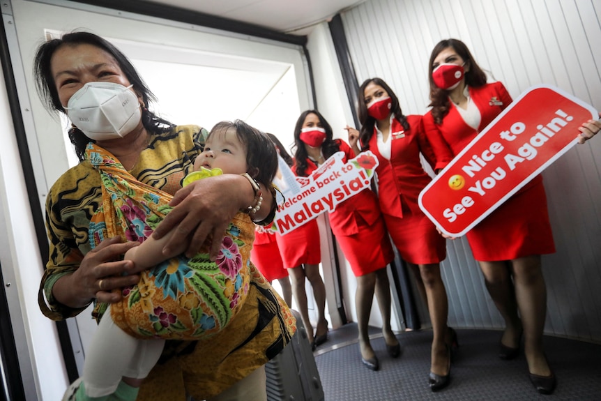 A woman smiling in a mask carrying a child with flight attendants in the background wearing red holding welcome signs