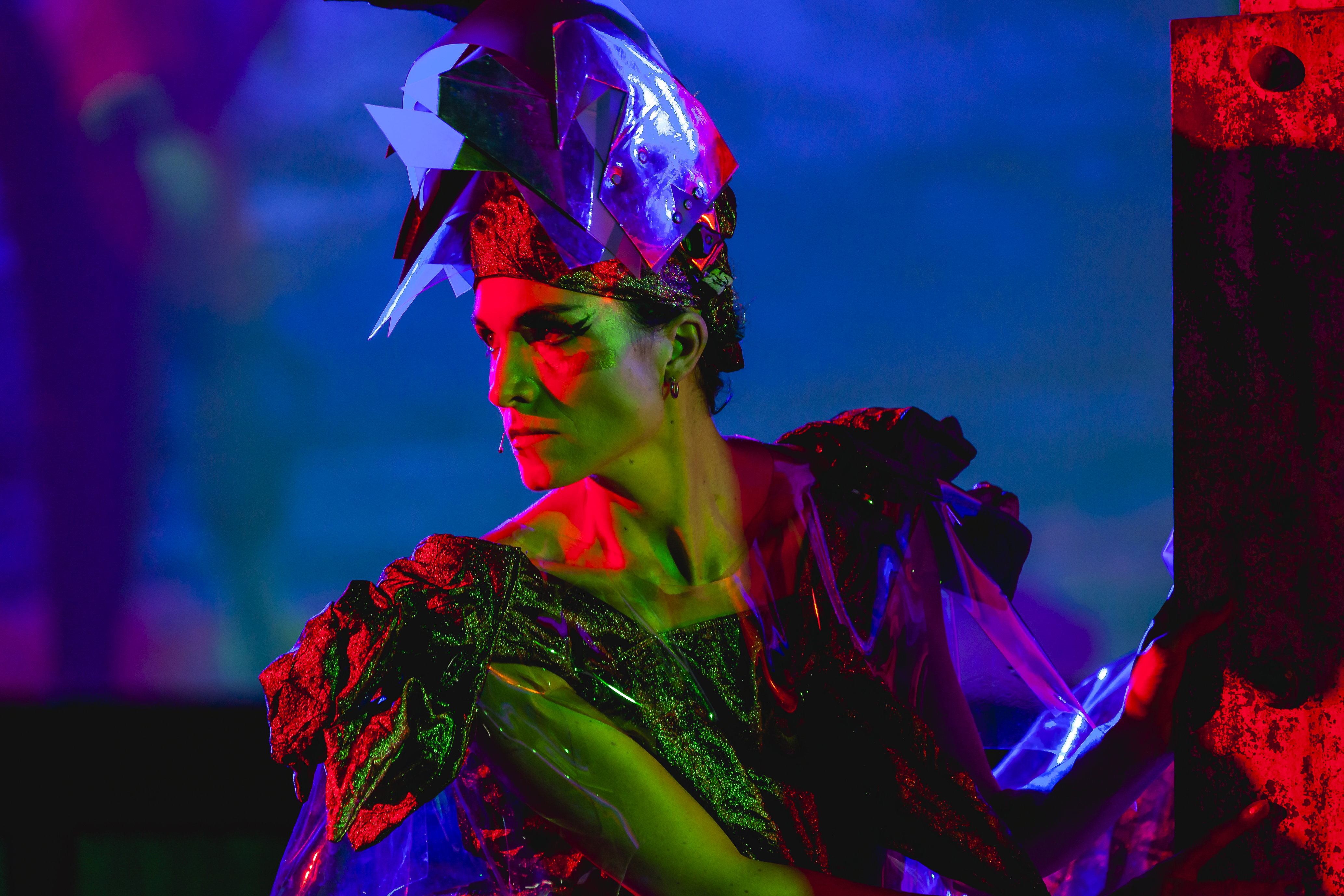 A person with a severe expression, looking to the side, wearing an ostentatious headdress and matching dress of futuristic blue.