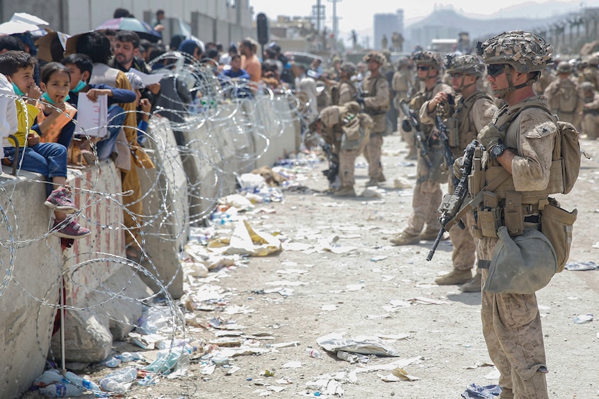 Afghans (left) stand in front of a wall, barbed wire and armed US soldiers